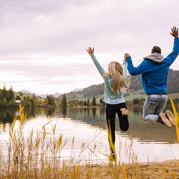 Ein Paar springt Hand in Hand fröhlich am Ufer eines ruhigen Sees in die Luft.